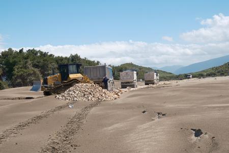 A road is being built in the sand; numerous lorries and a bulldozer can be seen.