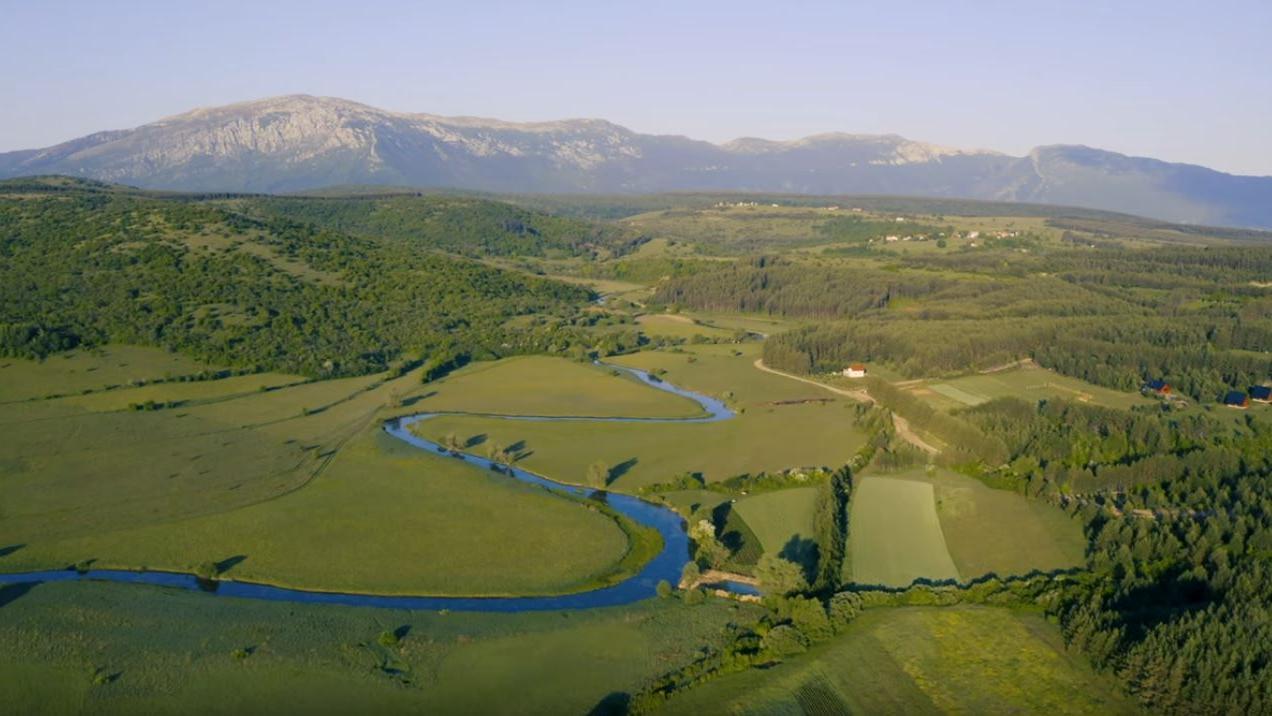 Screenshot: Ein Fluss schlängelt sich durch ein Tal mit Wiesen und Feldern.