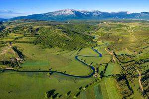 Lufbild des Livanjsko Polje mit grünen Wiesen einem Fluss und den Bergen im Hintergrund.