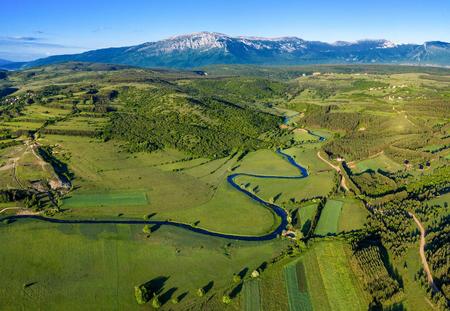Lufbild des Livanjsko Polje mit grünen Wiesen einem Fluss und den Bergen im Hintergrund.