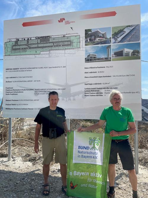 Gabriel Schwaderer and Richard Mergner in front of construction site poster at Vlora Airport