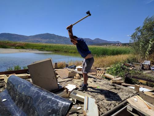 A man standing on the shore of a lake among rubble, smashing a cupboard with an axe