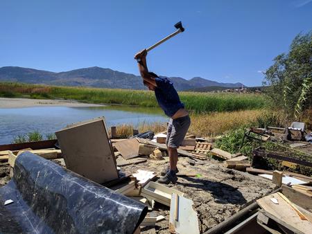 A man standing on the shore of a lake among rubble, smashing a cupboard with an axe