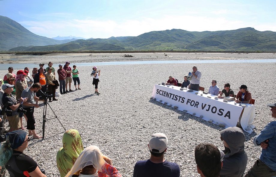 Mehrere Menschen sitzen an einem langen Tisch auf einer Flussinsel der Vjosa und geben eine Pressekonferenz.